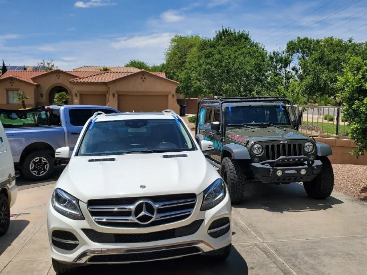 Alt text: A Mercedes-Benz SUV parked in front, with a Jeep Wrangler and a truck in a sunny driveway surrounded by greenery.