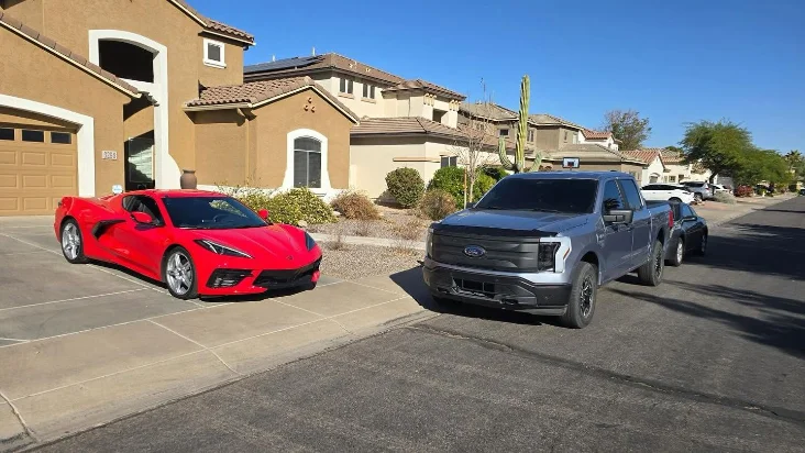 Red Corvette and gray Ford electric truck parked in a suburban neighborhood on a sunny day. Beautiful homes in background.